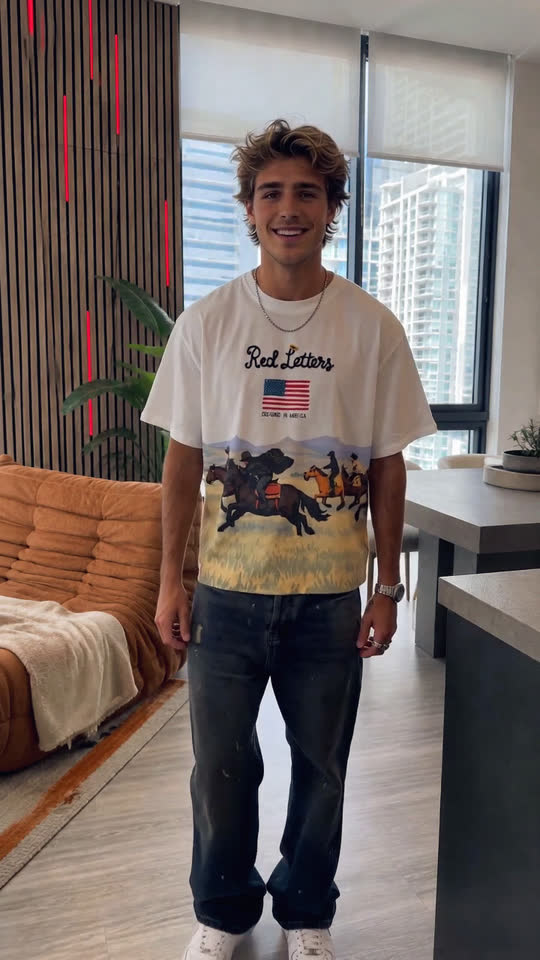 A man wearing an oversized printed graphic t-shirt and dark jeans turning and walking through a bright apartment interior, showing the front and back of the print.